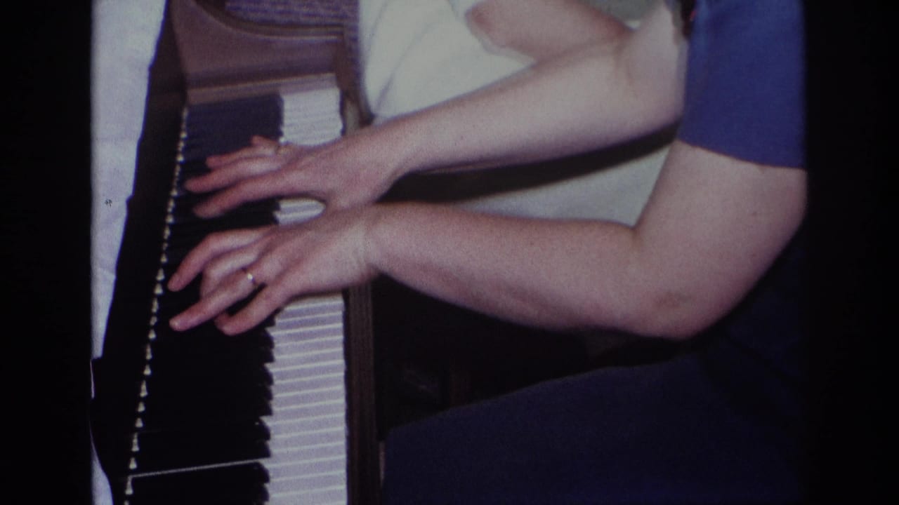 Close-up 1967 8mm frame from Rockaway Beach showing a person's hands on piano keys, with short-sleeved clothing and dark piano trim visible.