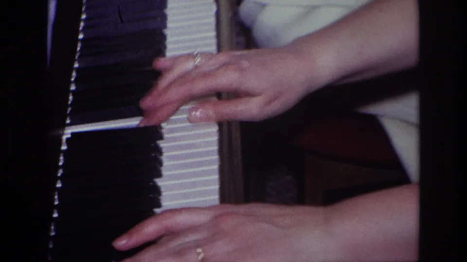 Close-up 1967 home-movie frame showing hands with rings moving across piano keys inside a private domestic setting.