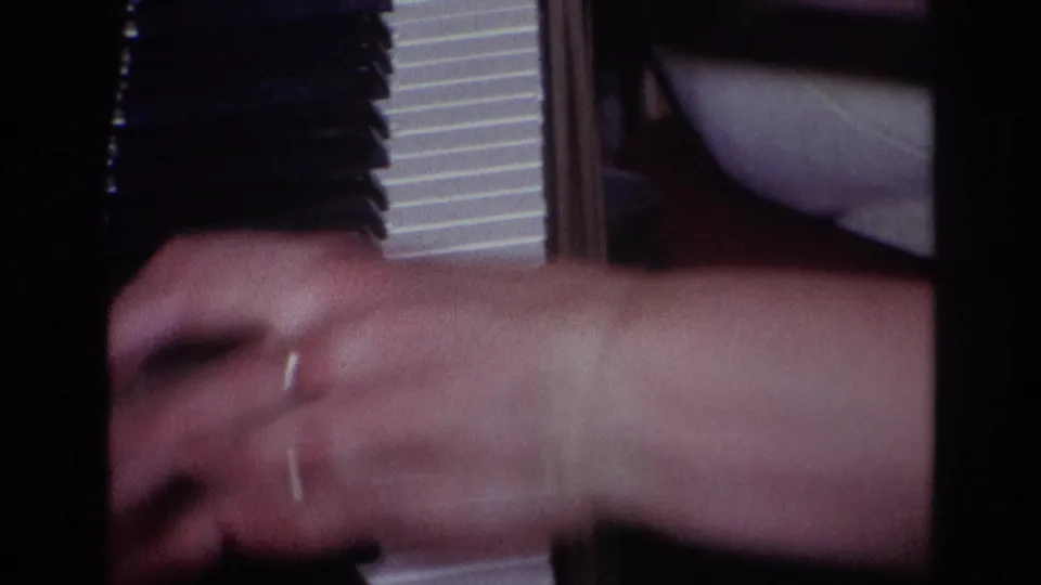 Motion-blurred 8mm frame of a hand passing across the piano keyboard, with a partial seated figure and interior furniture visible.