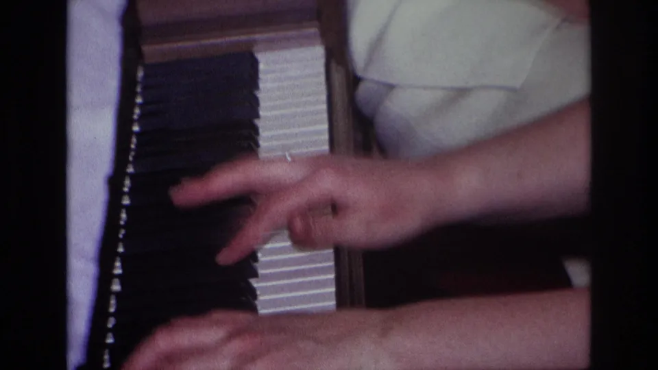 Final close-up frame from the 1967 Rockaway Beach home movie showing hands on the piano keys, rings on the fingers, and a light garment in the background.
