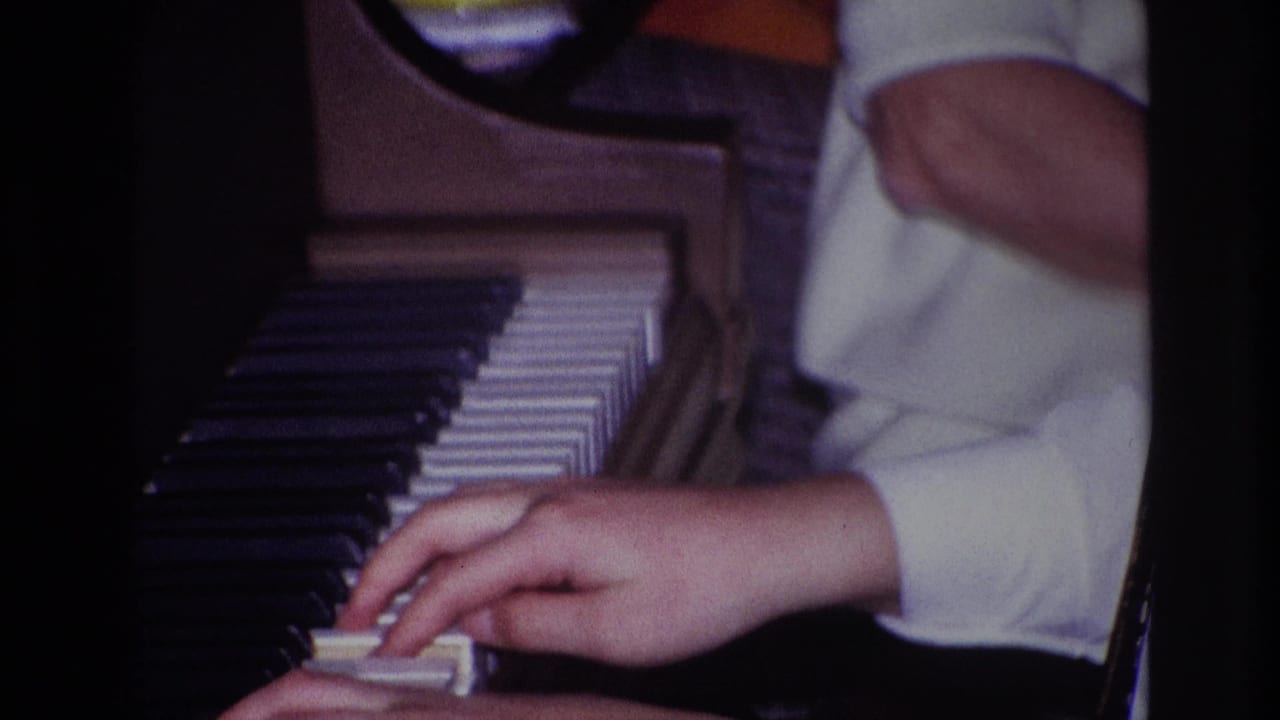 Close-up from 1967 Rockaway Beach home-movie footage showing hands in a white sleeve playing black and white piano keys indoors.