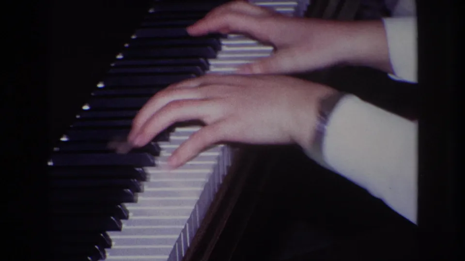 Close-up 8mm frame of a person playing piano, with both hands, a white sleeve, and a wristwatch visible near the dark wooden instrument.