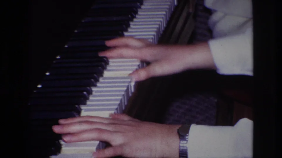 Final close-up frame from the 1967 Rockaway Beach home movie showing hands on piano keys, a wristwatch, and a dark interior piano surface.