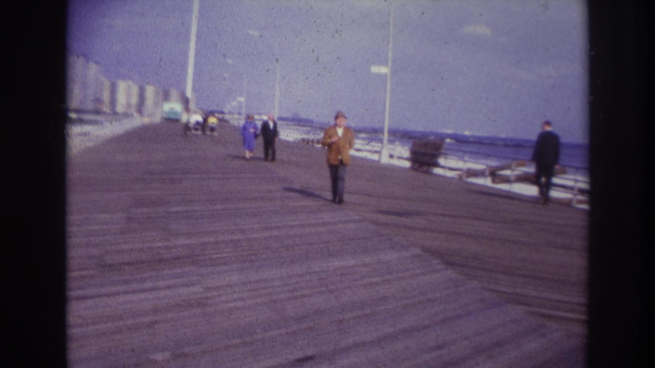 1967 Rockaway Beach home-movie frame showing a man in a brown jacket walking toward the camera on a boardwalk near the ocean.