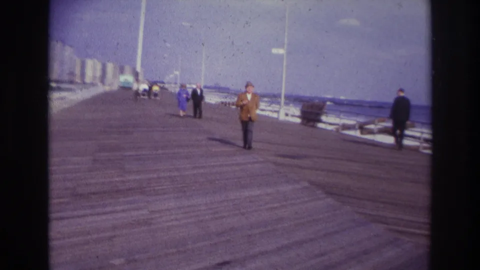 1967 Rockaway Beach home-movie frame showing a man in a brown jacket walking toward the camera on a boardwalk near the ocean.