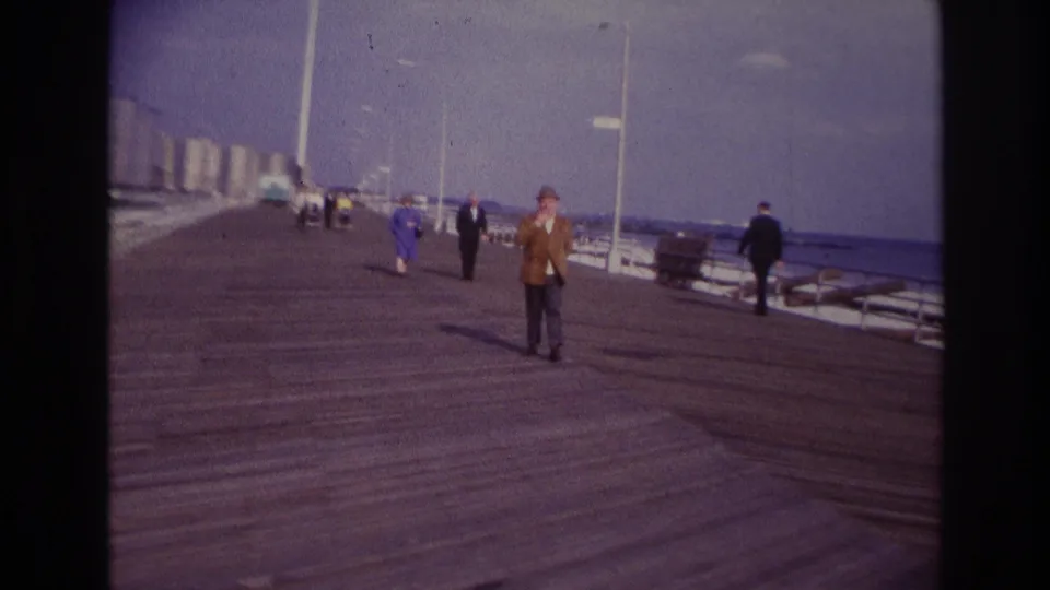 1967 Rockaway Beach boardwalk frame showing a man smoking as he passes benches and distant pedestrians near the ocean.