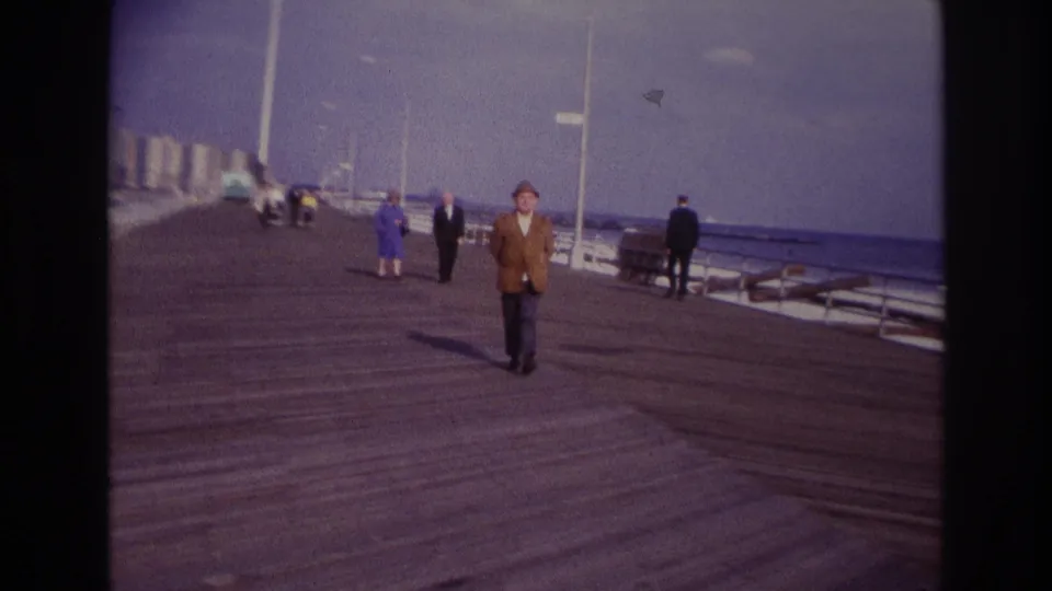 Rockaway Beach home-movie frame from 1967 with the smoking man closer to camera on the wooden boardwalk.