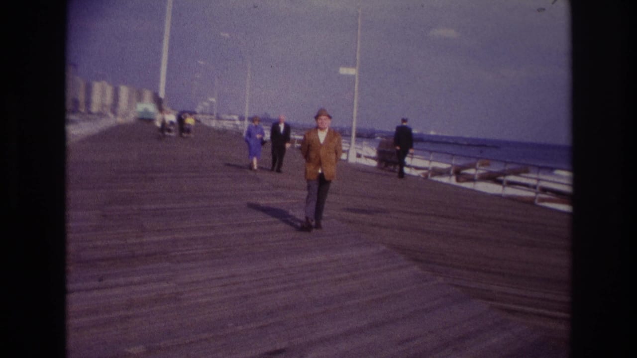1967 Rockaway Beach home-movie frame showing a dressed man in a hat walking along the boardwalk near benches, railings, and distant visitors.