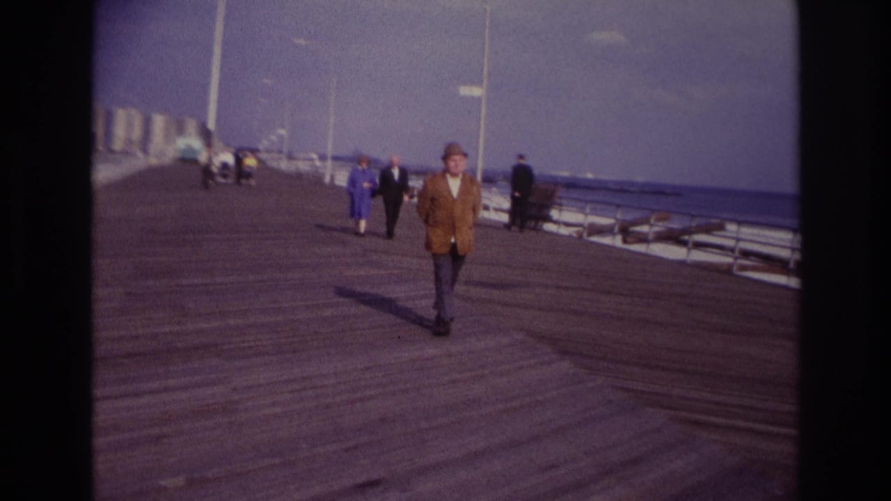 Man in hat continues walking on the boardwalk as people stroll in the background near the ocean side.