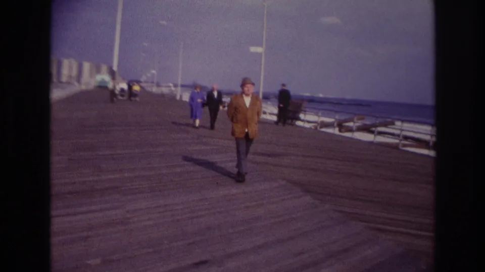 1967 Rockaway Beach frame with the central man walking closer to camera on a broad wooden boardwalk, with benches and seaside railing to the right.