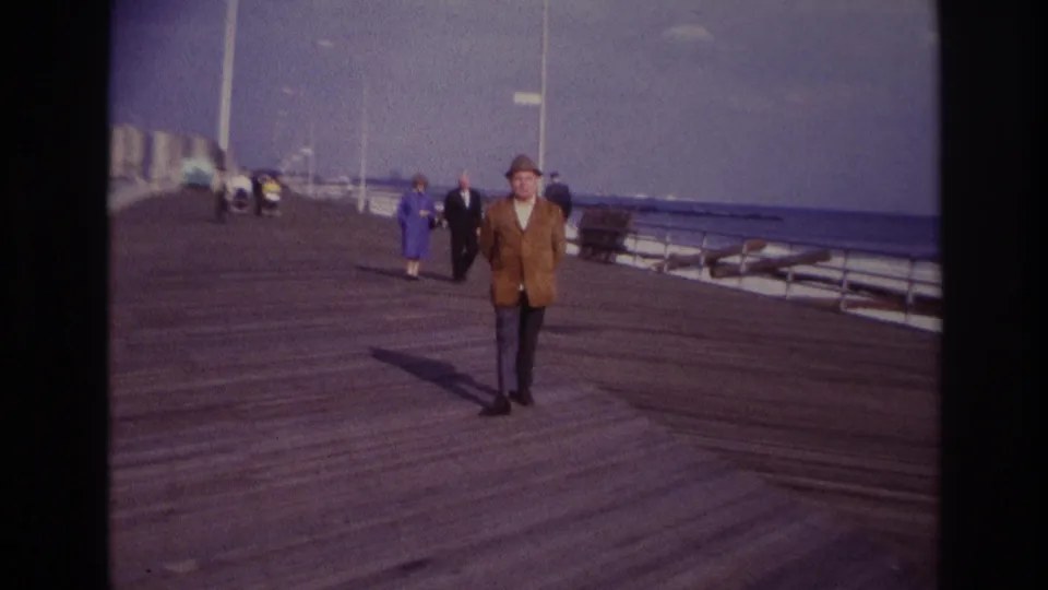 Rockaway Beach, 1967 home-movie frame showing the man closer in view, with a woman in blue and other pedestrians behind him on the promenade.