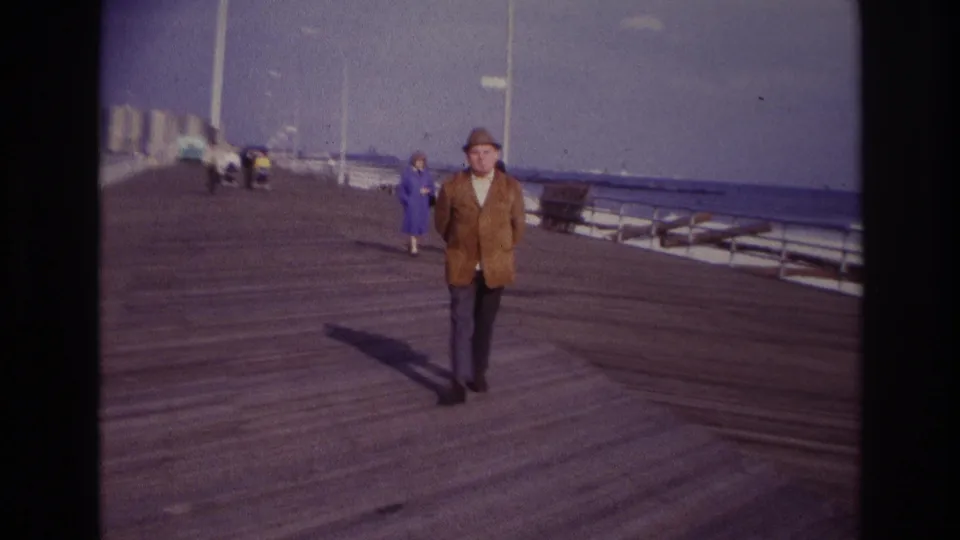 Final 1967 Rockaway Beach frame with the man near the camera, standing or slowing on the boardwalk as background visitors and benches remain visible.