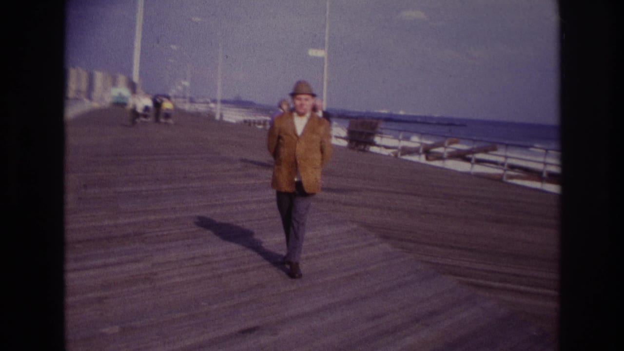 At Rockaway Beach in 1967, a man in a brown jacket and fedora approaches the camera on a wooden boardwalk near calm ocean water.