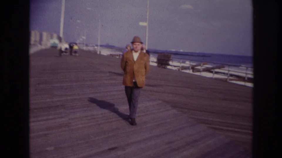 At Rockaway Beach in 1967, a man in a brown jacket and fedora approaches the camera on a wooden boardwalk near calm ocean water.