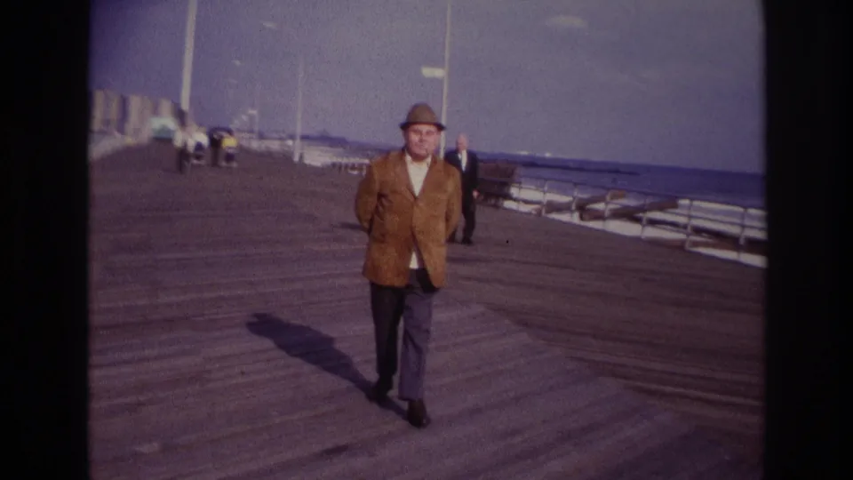 The Rockaway Beach boardwalk scene continues as the man nears the camera, with the ocean and a few background visitors visible.
