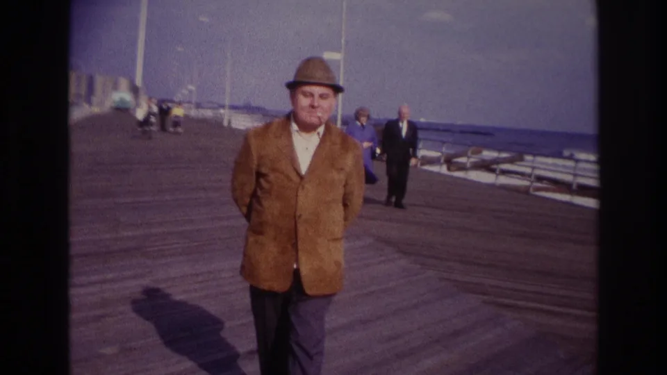 The man reaches a closer position on the Rockaway Beach boardwalk, with background pedestrians and calm ocean water still visible.
