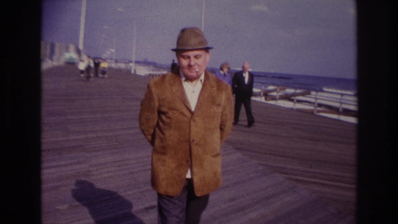 At Rockaway Beach in 1967, a man in a brown jacket and fedora walks toward the camera on a wooden boardwalk with the ocean behind him.