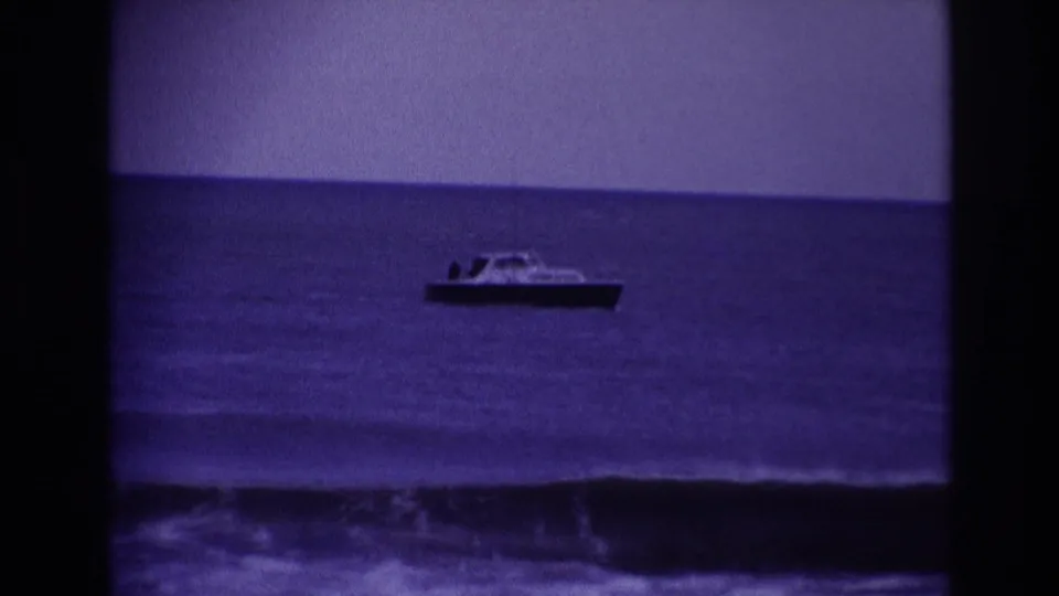 A small boat appears offshore at Rockaway Beach in 1967, with dark blue water, a clear horizon, and low surf near the bottom of frame.