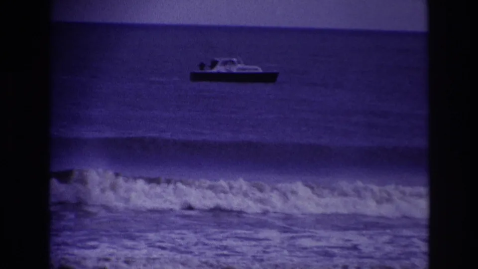 1967 Rockaway Beach 8mm frame with a distant small motorboat on open water and surf breaking across the foreground.