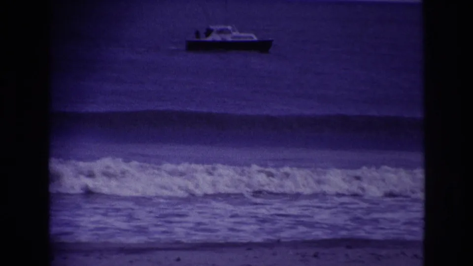 1967 Rockaway Beach 8mm frame showing a small offshore boat above a broad swell and foamy surf in the foreground.