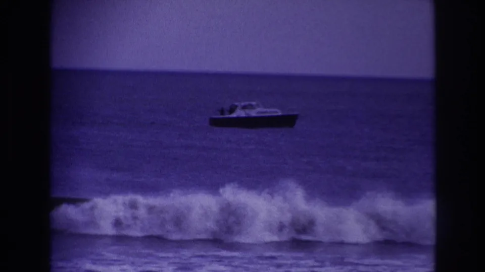 1967 Rockaway Beach frame with a small motorboat beyond a breaking wave, tinted blue by the 8mm film scan.
