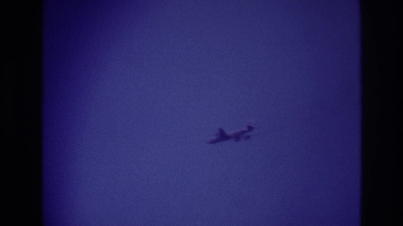 Small airplane crossing the sky in 1967 Rockaway Beach home-movie footage, seen as a soft silhouette against blue-purple 8mm film color.