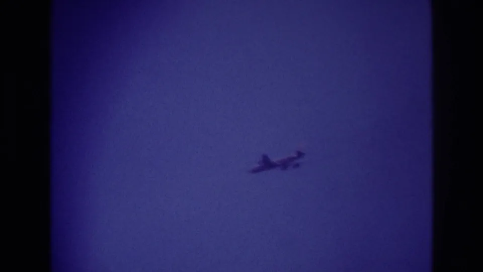 Small airplane crossing the sky in 1967 Rockaway Beach home-movie footage, seen as a soft silhouette against blue-purple 8mm film color.