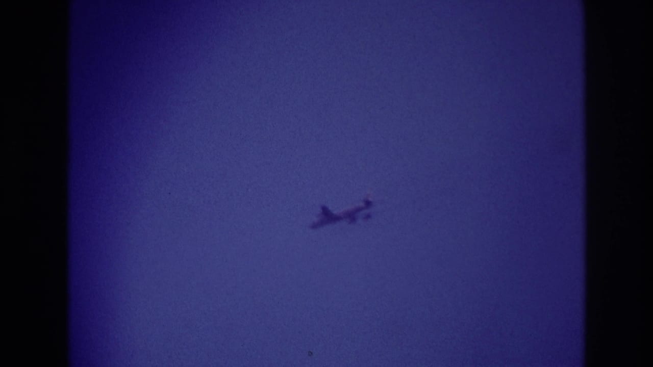 Small aircraft remains high against the blue sky in the Rockaway Beach 1967 8mm clip, with grain and faded color visible.