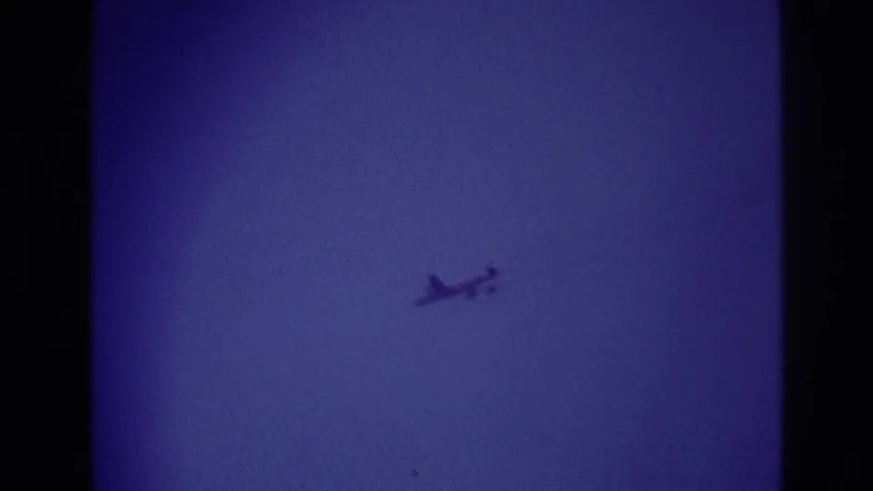 Small aircraft remains high against the blue sky in the Rockaway Beach 1967 8mm clip, with grain and faded color visible.