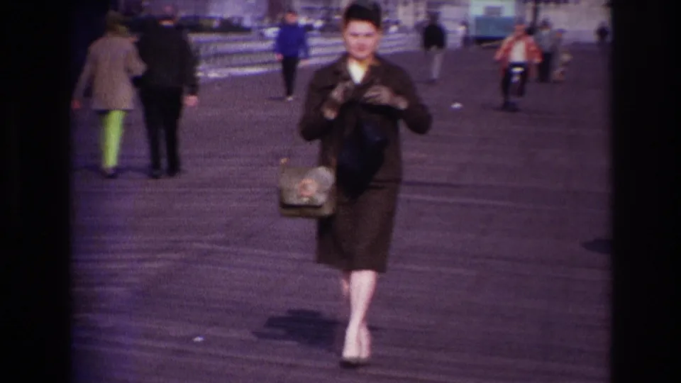 A woman in a dark suit and heels walks along the Rockaway Beach boardwalk in 1967, carrying a handbag as pedestrians move behind her.