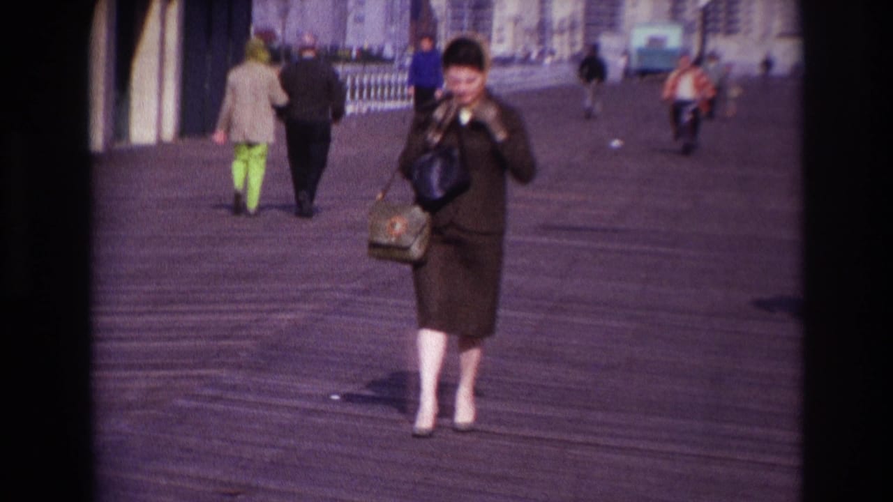 Woman in a dark dress and sunglasses walks on a wooden boardwalk while touching her hat.