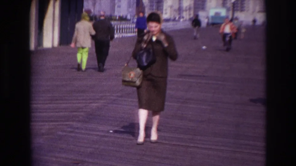 In 1967 Rockaway Beach, a woman in sunglasses walks toward the camera on the boardwalk while adjusting her hat.