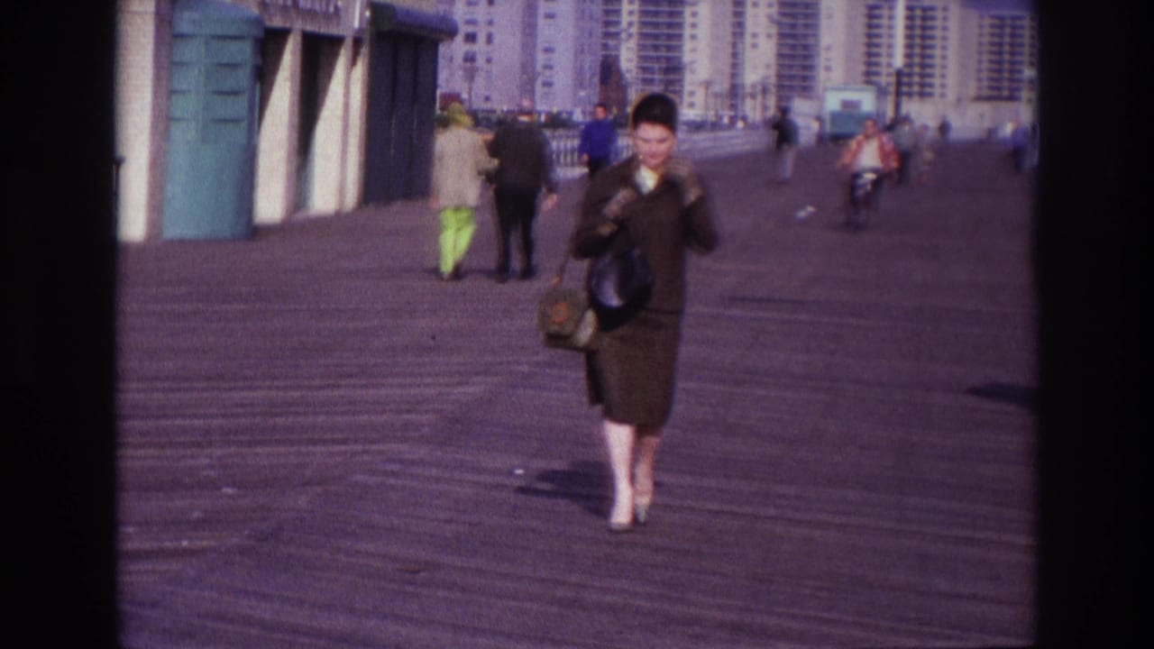Woman continues along the boardwalk with pedestrians and apartment buildings behind her.