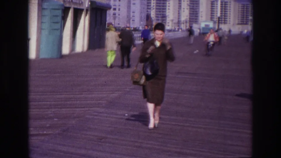 The woman walks farther along the Rockaway Beach boardwalk, with pedestrians and tall residential buildings in the background.