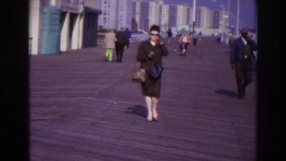 A woman in 1960s dress and sunglasses walks on the Rockaway Beach boardwalk while another adult pedestrian passes at right.