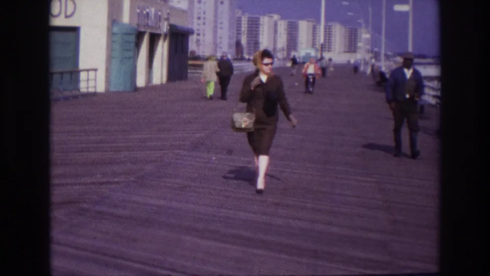 The woman moves across the Rockaway Beach boardwalk near the end of the shot, with passersby and beachfront buildings behind her.