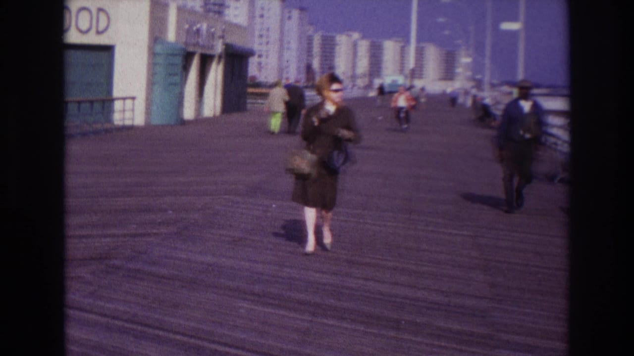 1967 Rockaway Beach boardwalk view with a woman in a dark coat approaching the camera near food storefronts and distant high-rises.