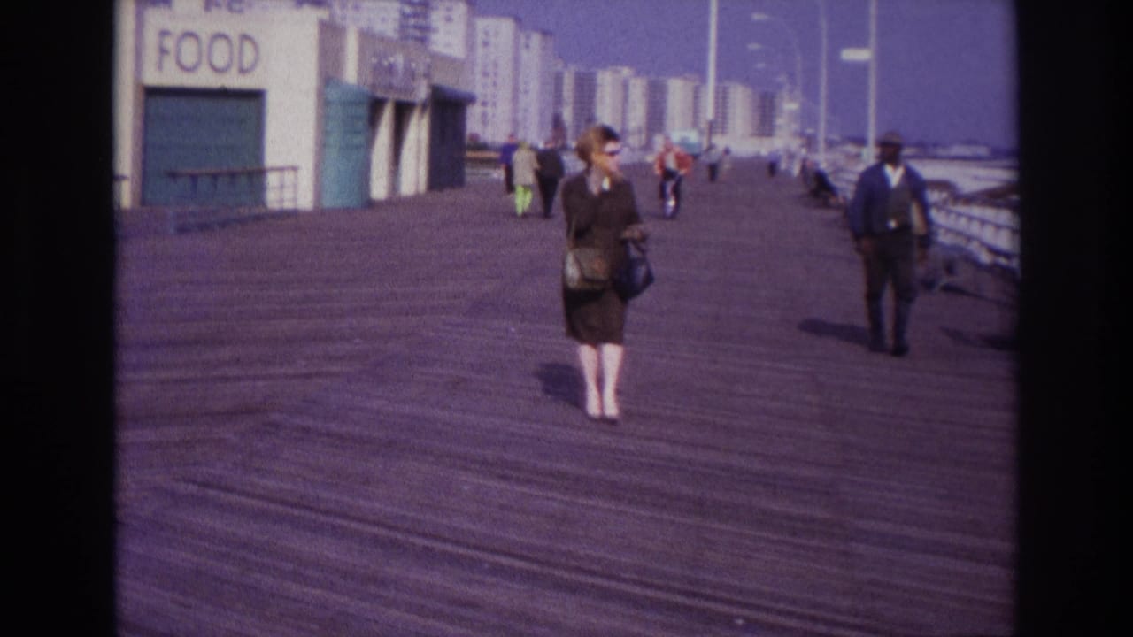 Rockaway Beach boardwalk in 1967 with a woman in sunglasses, a suited pedestrian, closed food stands, railings, and apartment blocks beyond.