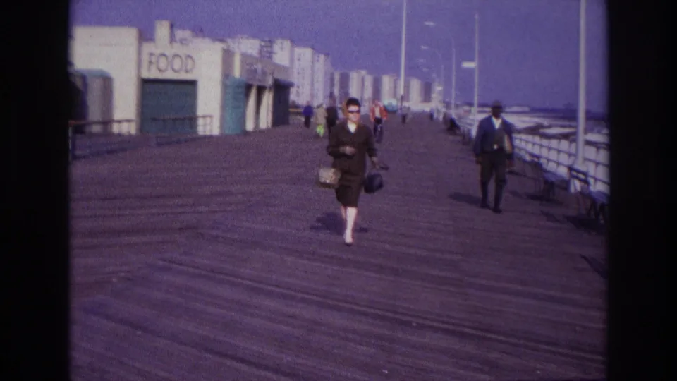 1967 Rockaway Beach boardwalk scene with a woman carrying a handbag, pedestrians behind her, food signage, railings, and hazy high-rises.