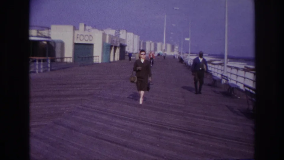 Wide 1967 Rockaway Beach boardwalk frame with a woman at center, suited man at right, food storefronts at left, railings, and skyline beyond.