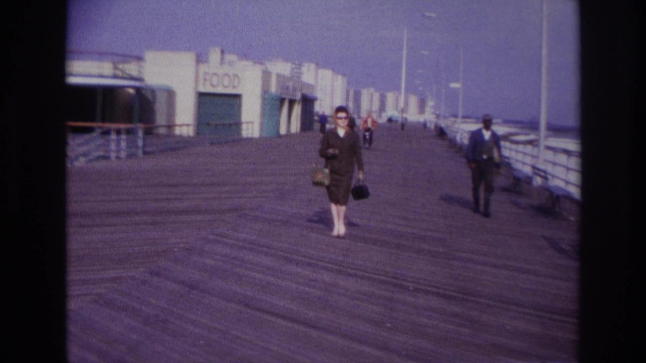 At Rockaway Beach in 1967, a woman in a brown suit and sunglasses walks along a wooden boardwalk near a FOOD sign and railings.