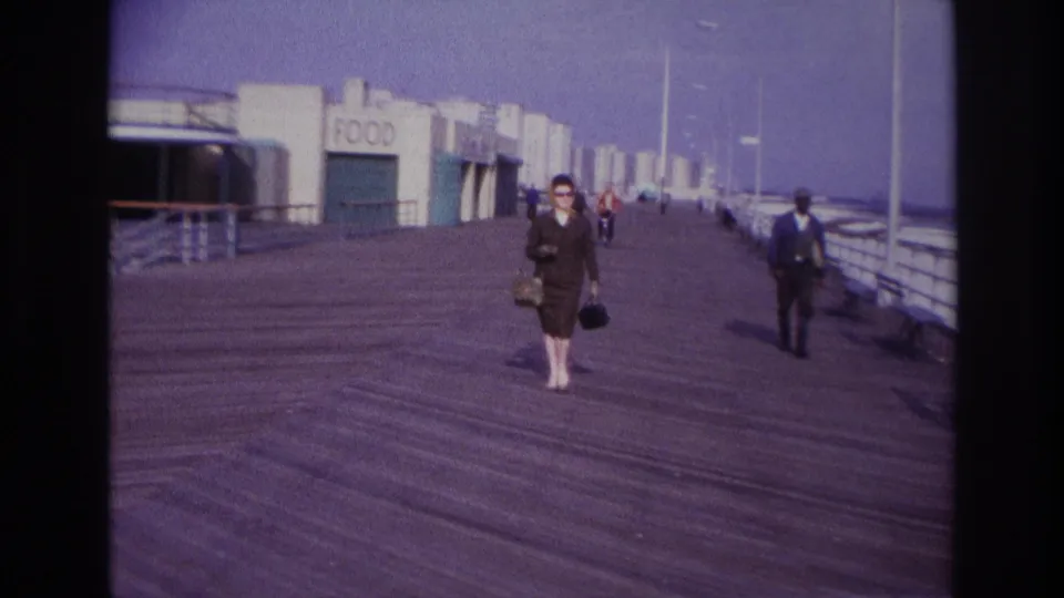 At Rockaway Beach in 1967, a woman in a brown suit and sunglasses walks along a wooden boardwalk near a FOOD sign and railings.
