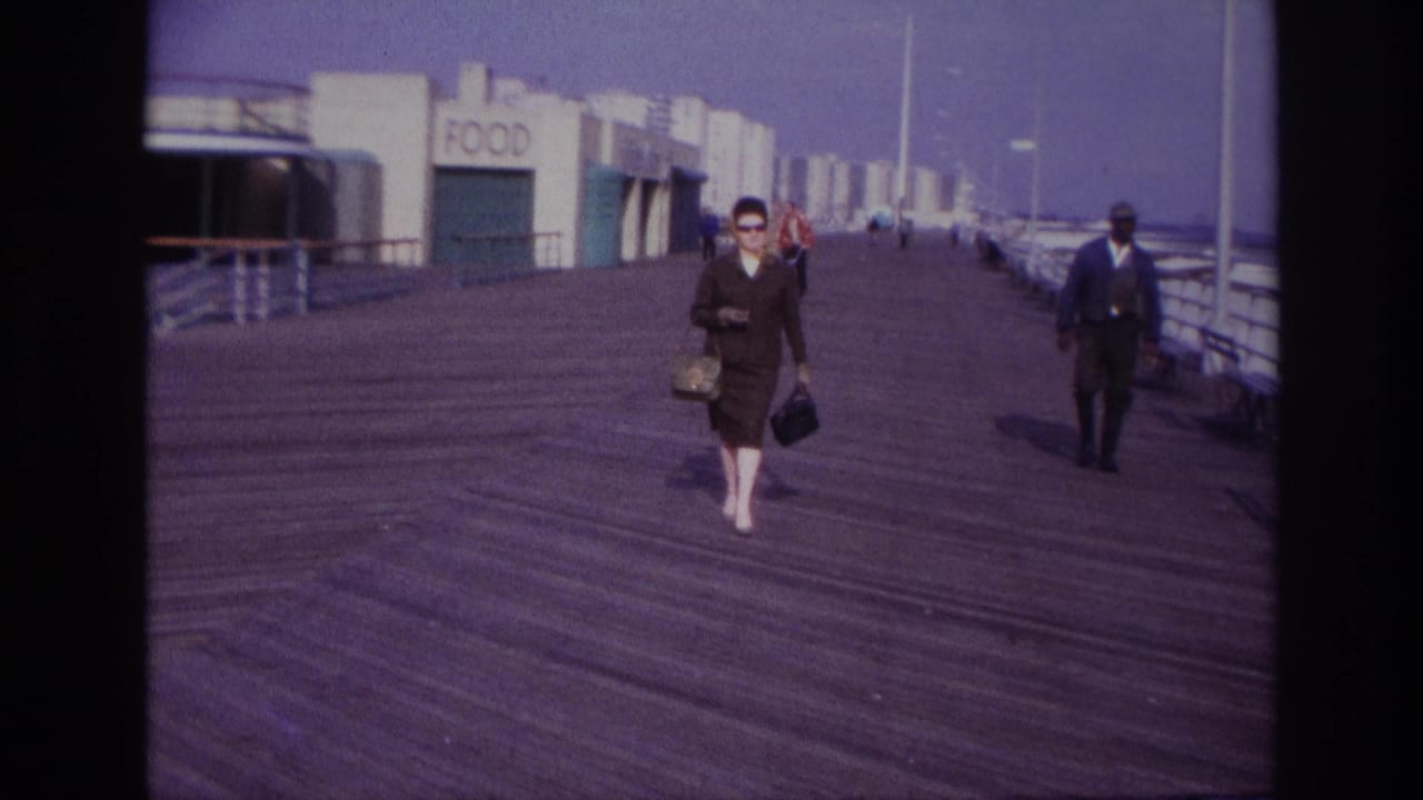 Woman with handbags walks closer on the boardwalk while a man in a dark suit passes at right.