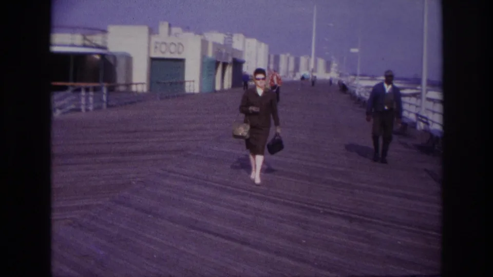 The woman continues toward the camera on the Rockaway Beach boardwalk, with a suited man, distant walkers, railings, and food buildings visible.