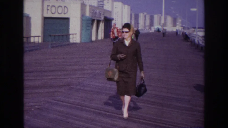 A closer 1967 Rockaway Beach boardwalk view shows the woman in a brown suit carrying two bags, with the FOOD sign and pedestrians behind her.