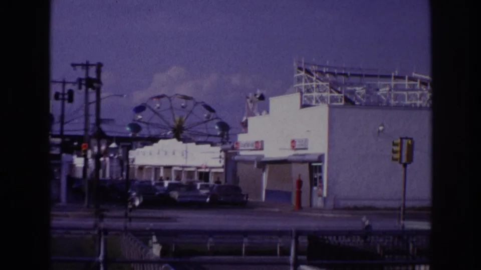 Wider 1967 Rockaway Beach boardwalk view with a Ferris wheel, concession building, parked cars, utility poles, and traffic signal.