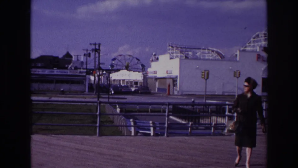 1967 Rockaway Beach boardwalk scene with a woman in a dark dress walking past railings, traffic lights, and distant amusement rides.
