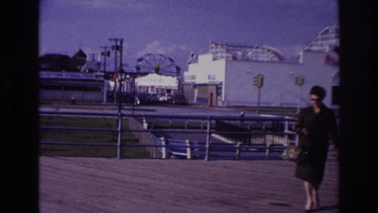 At Rockaway Beach in 1967, a woman in a dark suit and sunglasses walks along the boardwalk with Playland rides in the background.
