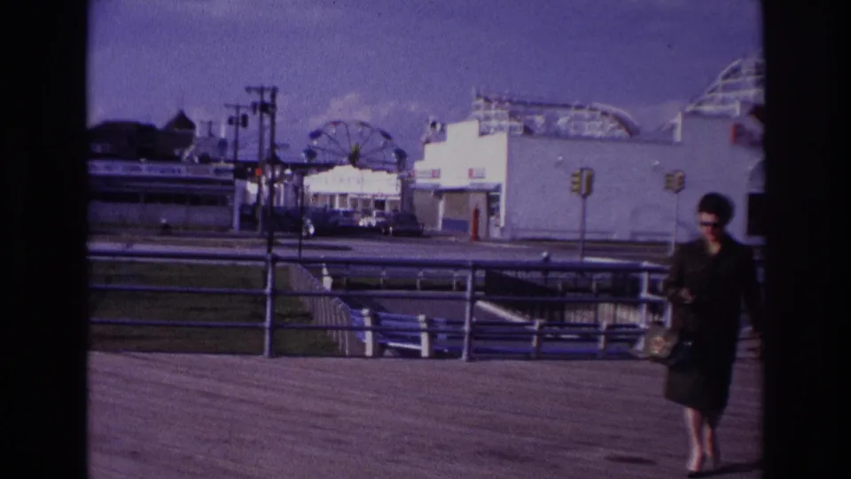 At Rockaway Beach in 1967, a woman in a dark suit and sunglasses walks along the boardwalk with Playland rides in the background.