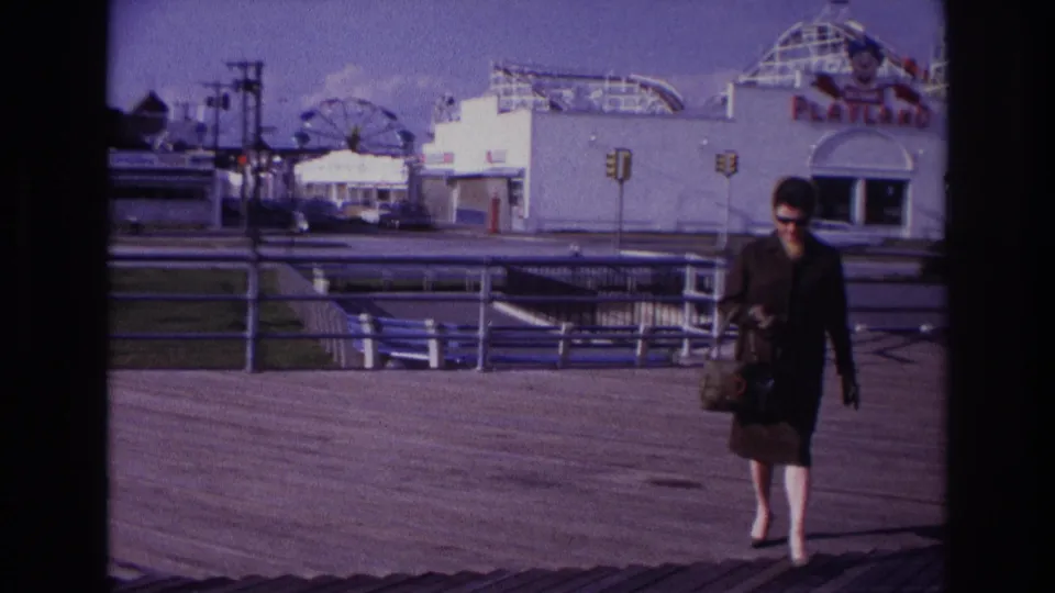 The woman continues across the Rockaway Beach boardwalk, carrying a handbag as the Playland building and rides remain visible behind her.